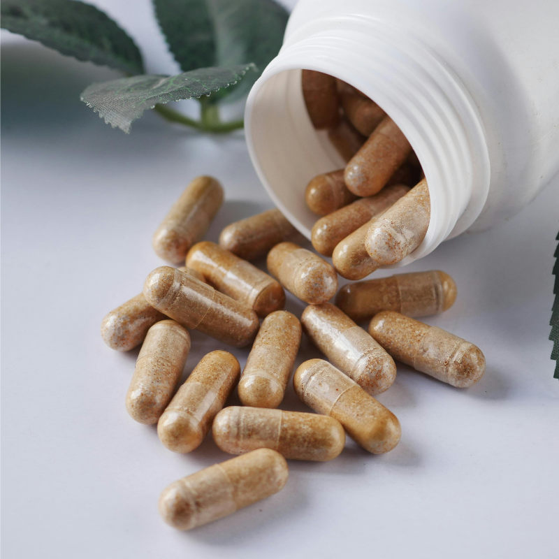 Capsules spilling out of a white container on a light surface with green leaves in the background
